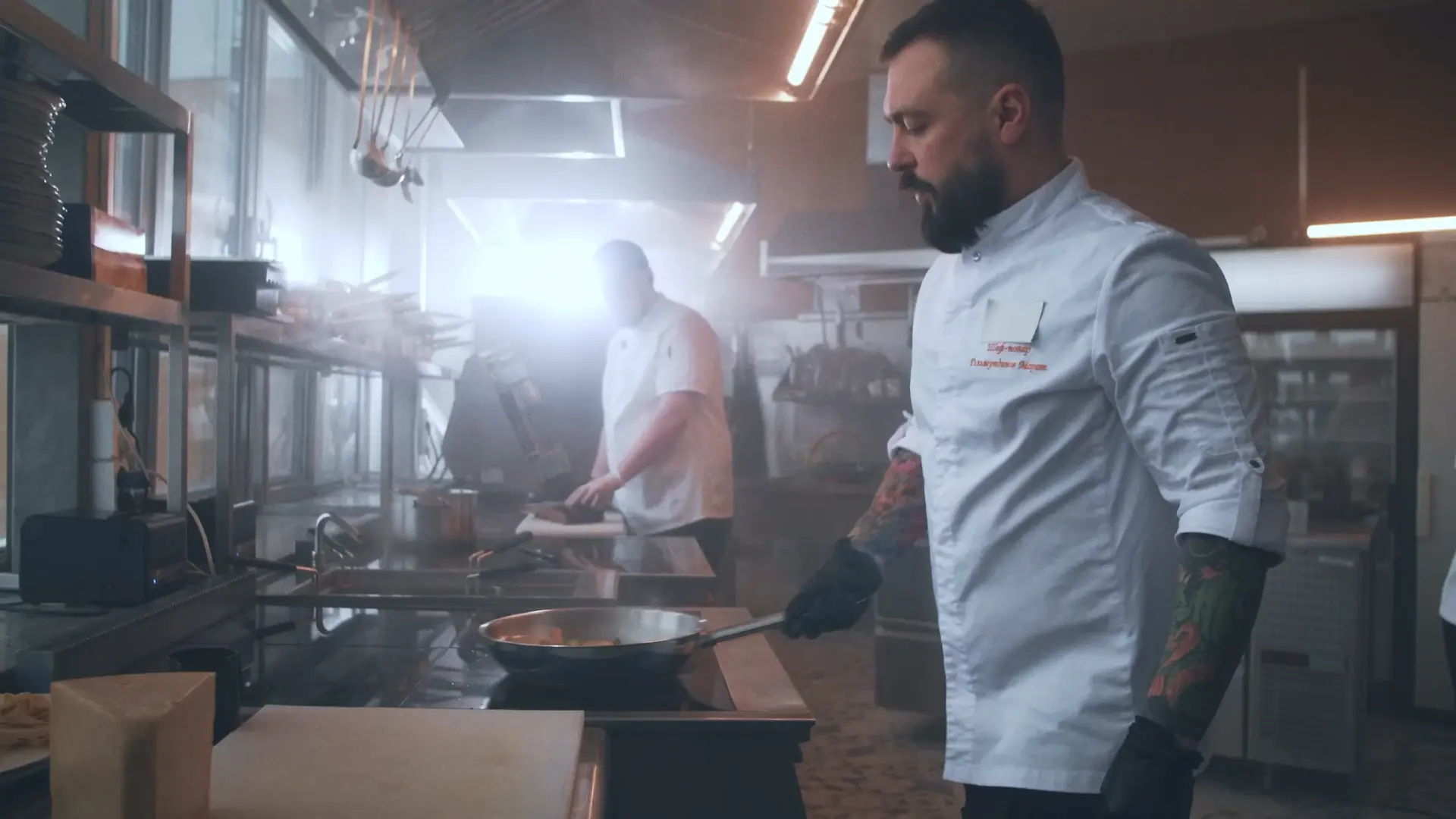 Kock lagar mat vid spis i ett professionellt restaurangkök medan en kollega arbetar i bakgrunden.